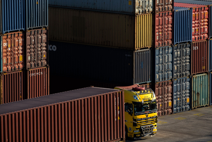 Close-up of truck maneuvering beside tall stacks of cargo containers at terminal.