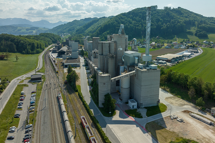 Rohrdorfer Werk mit Bahnanschluss inmitten der Landschaft; Güterzug auf den Gleisen neben dem Werk.